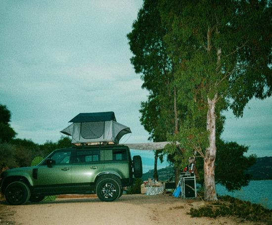 Prezzo del successo: Land Rover Defender verde con tenda da tetto parcheggiato vicino a un albero e al mare in Sardegna